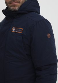 Navy blue insulated jacket with a hood, featuring an orange emblem and patches on the chest and sleeve. Smooth fabric with pockets.