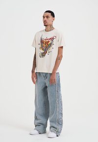 Béžové bavlněné tričko s barevným potiskem orla a výrazným textem. Volné světle modré džíny s roztrhanými detaily na bocích. Bílé boty.