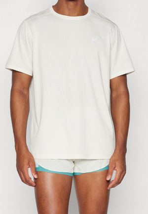 Mand iført en ensfarvet hvid kortærmet T-shirt og hvide atletikshorts med turquoise kant mod en neutral baggrund.