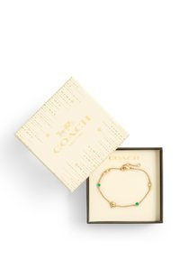 Gold chain bracelet with green stones and a star accent, displayed in a cream box featuring gold graphics and "COACH" branding.