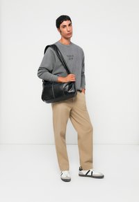 Ung mann iført grå hettegenser, beige bukser, hvite sneakers, holder en svart skinn skulderveske, står mot en hvit bakgrunn.