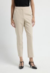 Beige op maat gemaakte broek met voorplooien, recht model en zijzakken, gecombineerd met zwarte hakken. Soepele stofstructuur.