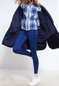 Modrá a bílá kostkovaná košile na knoflíky; tmavě modré přiléhavé džíny; bílé tenisky; tmavě modrý kabát částečně rozepnutý, odhalující vnitřní podšívku.