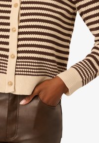 Hnědo-krémový pruhovaný cardigan s texturovaným pleteným vzorem, se zlatými knoflíky a žebrovanými manžetami, nošený přes hnědé kožené kalhoty.