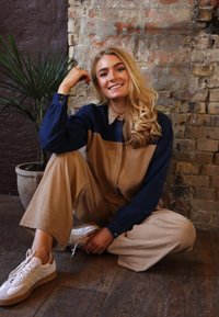 Kordbársony ruha kéttónusú kialakítással: sötétkék fent és bézs alul. Fehér tornacipők bézs részletekkel, egy texturált fal előtt.