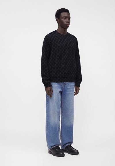 Homme debout portant une chemise noire à manches longues à motifs, un jean large bleu clair et des chaussures noires sur un fond clair uni.