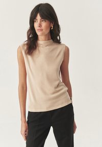 Beige bezramenný top s vysokým stojáčkem, vyrobený z hladké tkaniny. Má přiléhavý střih a jemnou elasticitu, kombinovaný s černými kalhotami.