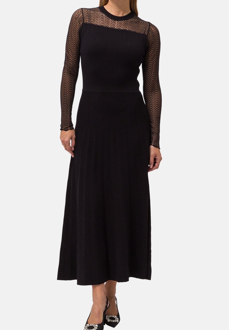Femme portant une longue robe noire avec des manches longues transparentes à motifs et une jupe plissée, associée à des talons noirs ornés.