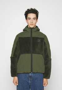 Veste matelassée verte avec un panneau en texture peluche, fermeture éclair à l'avant et capuche. Elle dispose de grandes poches latérales et d'une finition noire pour le contraste.