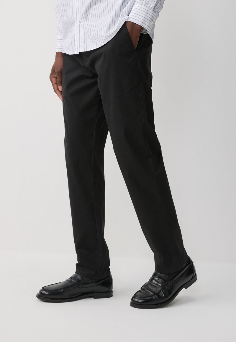 Czarne spodnie o prostym kroju, wykonane z gładkiej tkaniny. Noszone z czarnymi mokasynami i białą koszulą w paski, prezentujące klasyczny styl.