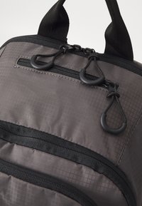 Sac à dos en tissu gris avec une surface texturée, doté d'une poche zippée à l'avant et d'accents en métal noir, y compris des cordons et des boucles réglables.