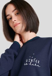 Sudadera azul marino con cuello redondo y acentos verdes. Presenta texto bordado "EL INVIERNO ha llegado" con puntos decorativos encima.