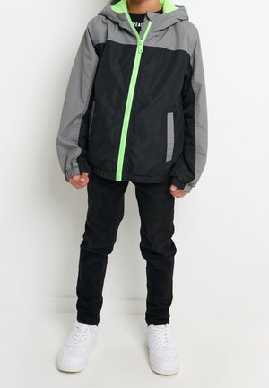 Sort og grå vindjakke med en grøn lynlås og hætte, udstyret med elastiske manchetter og sidelommer, parret med sorte bukser og hvide sneakers.