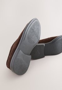 Chaussures en daim marron avec une semelle en caoutchouc grise flexible, présentant des accents texturés et une coque de talon ronde, vues en plongée sous un angle.
