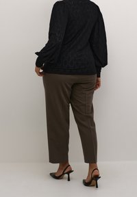 Personne portant un pantalon droit marron, des talons slingback noirs pointus et un haut noir texturé à manches longues, debout devant un fond gris uni.