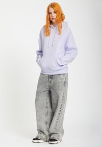 Lichtpaarse hoodie met een kangoeroezak, gecombineerd met hoog ingezette grijze wijdlopende jeans. Zwarte sneakers met witte accenten.