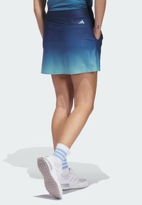 Blauwe gradient tennisrok met een aansluitende taille, twee zakken en een gestructureerd patroon, gecombineerd met witte sportschoenen en gestreepte sokken.