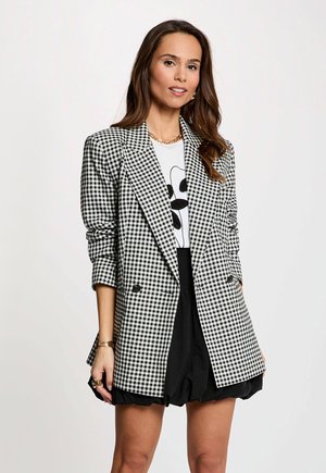 Femme portant un blazer à carreaux noir et blanc, un t-shirt graphique blanc, une jupe noire, un collier en or et un bracelet, posant devant un fond uni.