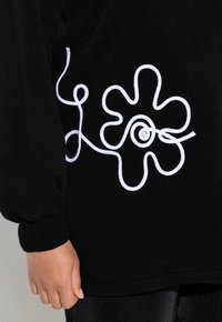 Schwarzer Pullover mit geripptem Textur und langen Ärmeln. Verfügt über ein weißes gesticktes Blumenmuster und geschlungene Akzente an der linken Seite.