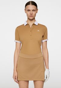 Vaaleanruskea polo-mekko, jossa on valkoinen kaulus ja hihansuut, vasemmassa rinnassa on logo. Mekko on yhdistetty tiukkaan hameeseen ja oikeassa kädessä on harmaa hansikas.