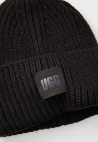Bonnet en tricot noir avec une texture côtelée et une étiquette noire cousue avec le logo "UGG" en gris foncé. Forme épaisse et ronde.