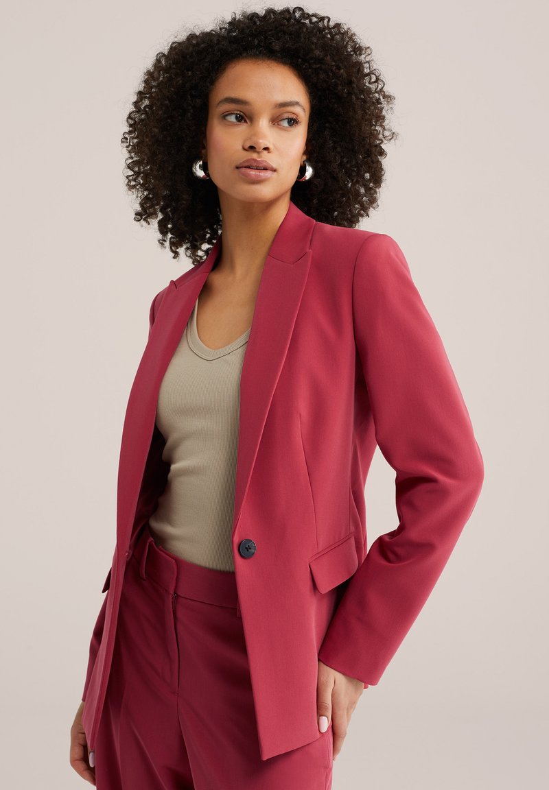Femme aux cheveux bouclés portant un blazer rose foncé et un pantalon assorti, accompagnés d'un haut beige à col en V et de boucles d'oreilles argentées en forme d'anneau, debout avec la main sur la hanche.