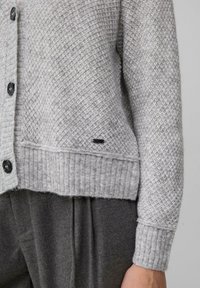 Graue strukturierte Strickjacke mit Knopfverschluss, geripptem Saum und Bündchen sowie einem kleinen schwarzen Akzentetikett. Getragen über dunkelgrauen Hosen.