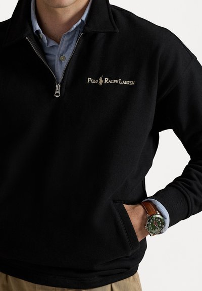 Hanorac negru cu fermoar, prevăzut cu guler, cu broderie aurie "Polo Ralph Lauren." Purtați peste o cămașă albă-albăstruie, asociat cu un ceas.