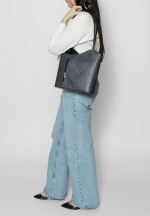 Borsa a tracolla con motivo struzzo in blu navy, chiusura con zip, manici neri e texture morbida, abbinata a jeans flared blu chiaro e scarpe nere a punta.