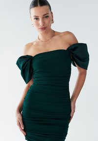 Ciemnozielona sukienka odkrywająca ramiona z bufkami na rękawach, marszczoną fakturą, dopasowanym krojem i subtelnym połyskiem. Gładka tkanina podkreśla kształt sylwetki.