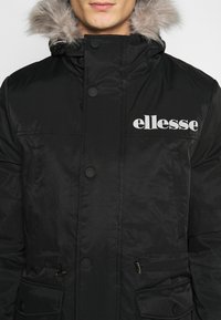 Parka negra con capucha forrada de piel, que presenta botones a presión, bolsillos con cremallera y el logo de "ellesse" en el pecho. Dise�ño elegante y resistente al agua.