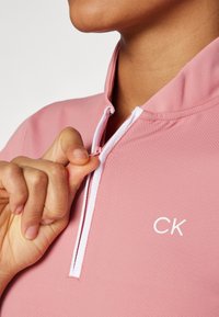Ροζ ζακέτα με φερμουάρ και υφή στο ύφασμα. Διαθέτει λευκό φερμουάρ και το λογότυπο "CK" σε λευκό χρώμα στην αριστερή πλευρά του στήθους.