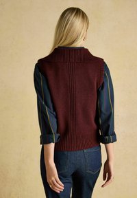 Burgunderfarbene Strick-Weste mit geripptem Kragen und Saum, die vertikale Nähte aufweist und mit einem gestreiften langärmeligen Hemd kombiniert ist.