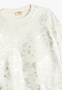 Felpa bianca con un motivo di glitter argento a macchie, scollo rotondo e polsini a costine. Tessuto morbido, design casual adatto per bambini.