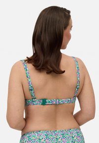 Sujeto colorido con un patrón, presenta un broche verde y tirantes ajustables. La tela muestra una mezcla de tonos púrpura, verde y rosa en un diseño floral.