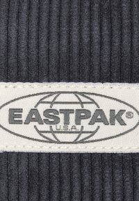 Tessuto nero a costine con un'etichetta beige che presenta il logo "EASTPAK" e un design del globo. Superficie texturizzata con dettagli di cucitura visibili.