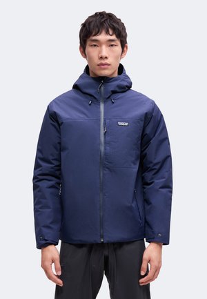 Uomo che indossa una giacca con cappuccio blu navy, con cerniera frontale e logo sul petto, in piedi davanti a uno sfondo chiaro e semplice.