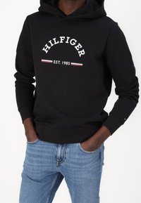 Felpa nera con cappuccio caratterizzata dal testo bianco "HILFIGER EST. 1985" e dettagli rosso-blu, con tasca a kanga e polsini a costine. Abbinata a jeans blu.