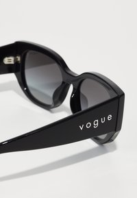 Black sunglasses with a thick frame, featuring dark lenses and a glossy finish. The arm displays the brand name "vogue" in white lettering.