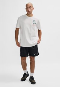 Vit kortärmad t-shirt med texten "OFF DUTY GEAR", kombinerad med svarta shorts som har "ATW" i grått. Svarta och olivfärgade sportskor.