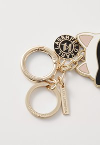 Portachiavi in tono oro con un design di gattino in stile cartone animato, dettagli neri e rosa, due anelli divisi e dettagli di branding incisi. Texture liscia.