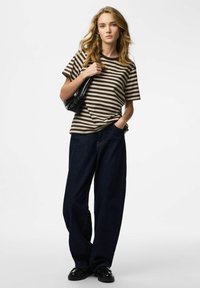 Prugasta pamučna majica u smeđoj i bež boji, uparena s širokim tamnim trapericama. Model drži crnu torbu i opušteno pozira.