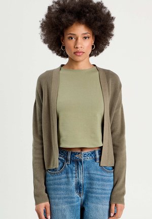 Olijfgroene cropped cardigan met ribbelstructuur, open voorkant en lange mouwen, gecombineerd met een aansluitend olijfgroen topje en blauwe high-waisted jeans.