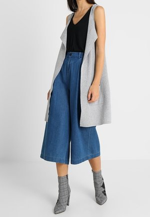 Kvinde iført ærmeløs grå cardigan, sort top, blå denim culotter med vide ben og grå mønstrede ankelstøvler, stående foran en hvid baggrund.