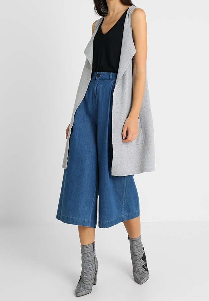 Femme portant un gilet sans manches gris, un haut noir, un pantalon culotte large en denim bleu et des bottines grises à motif, debout devant un fond blanc.