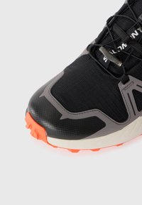 Black athletic shoe with textured mesh upper, grey accents, and bright orange outsole. Features a streamlined design and lacing system.
