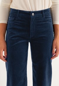 Navy blue corduroy pants feature a straight cut, two front pockets, a coin pocket, and a front button closure. Soft texture, smooth finish.
