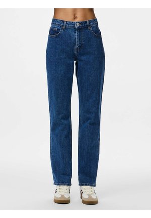 Personne portant un jean bleu taille haute à jambe droite et des baskets blanches, debout devant un fond clair et uni.