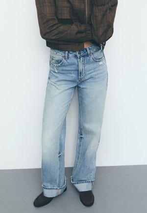 Lichtblauwe, wijde jeans met een vervaagde afwerking en een uitrafeling aan de onderkant. Beschikt over vijf zakken en een knoopsluiting. G gedragen met donkerbruine schoenen.