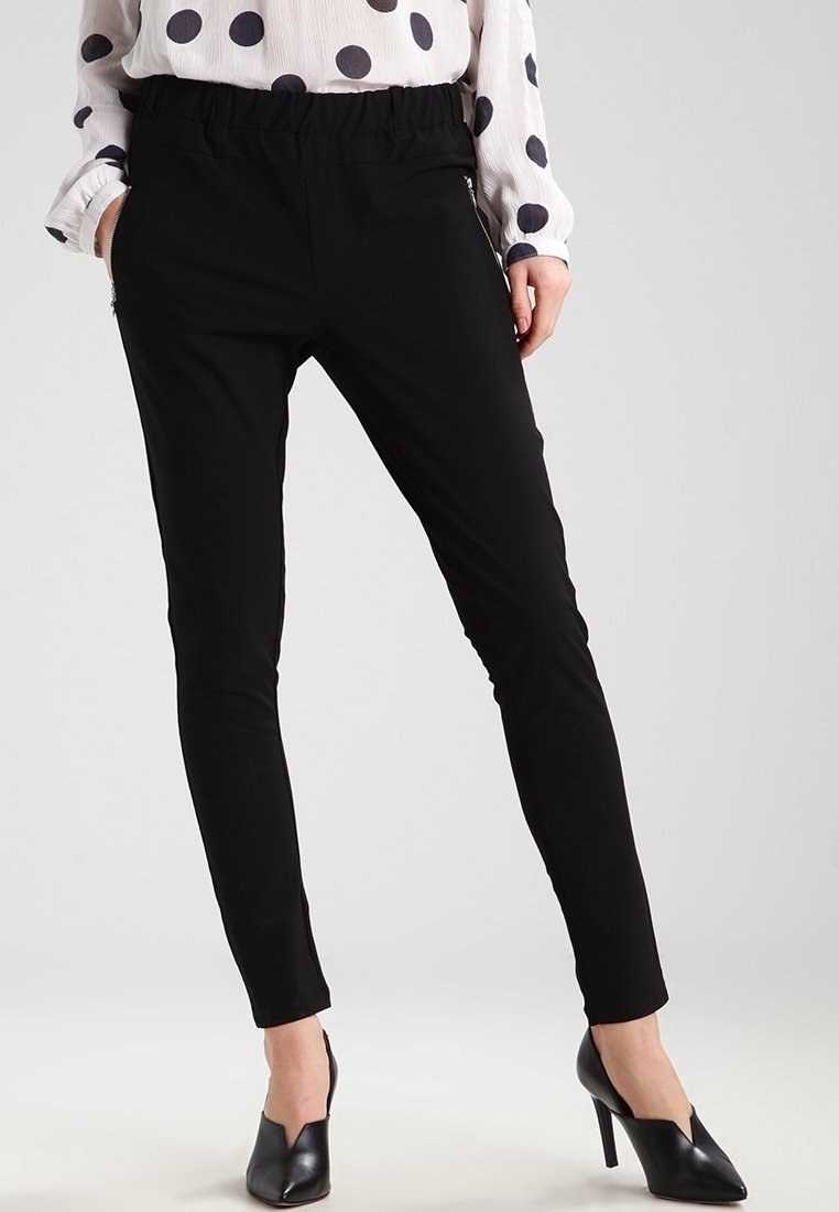 Femme portant un pantalon noir slim avec des fermetures éclair sur les poches, un chemisier blanc à pois noirs et des chaussures à talons hauts noires.